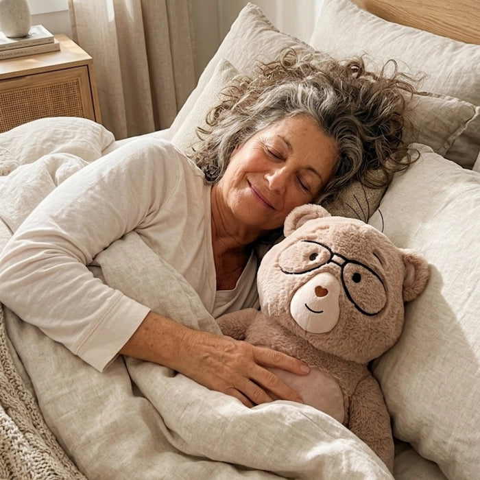 Woman lying in bed with a Percy bear wearing glasses, surrounded by soft bedding.