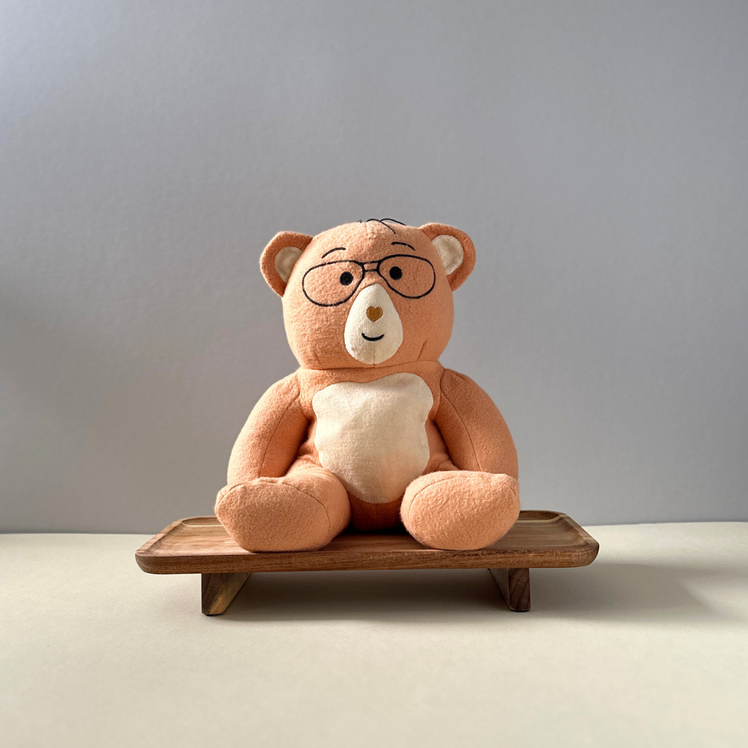 Plush teddy bear with glasses on a wooden stand against a plain background