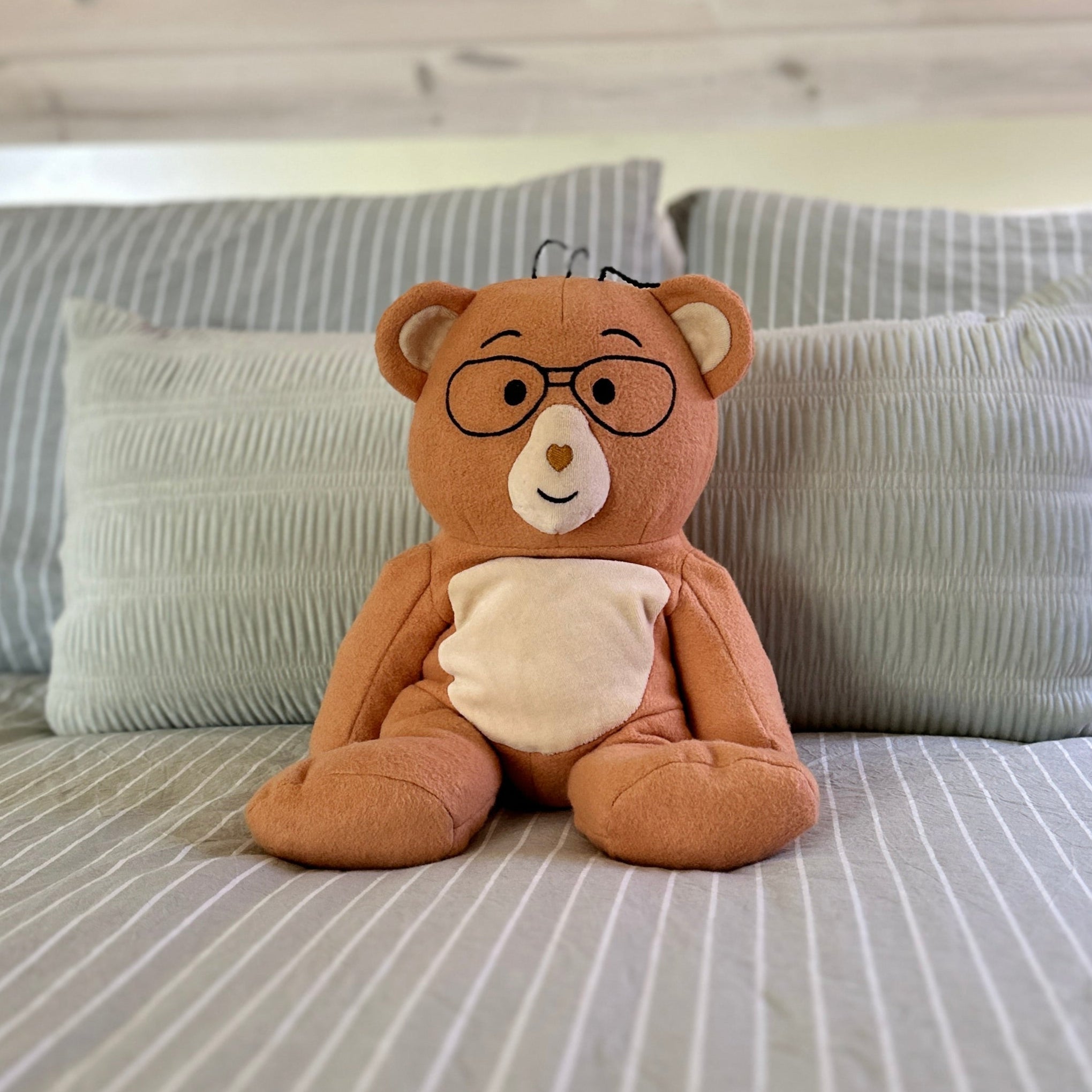 Plush teddy bear with glasses on a bed with striped bedding and decorative wall art.