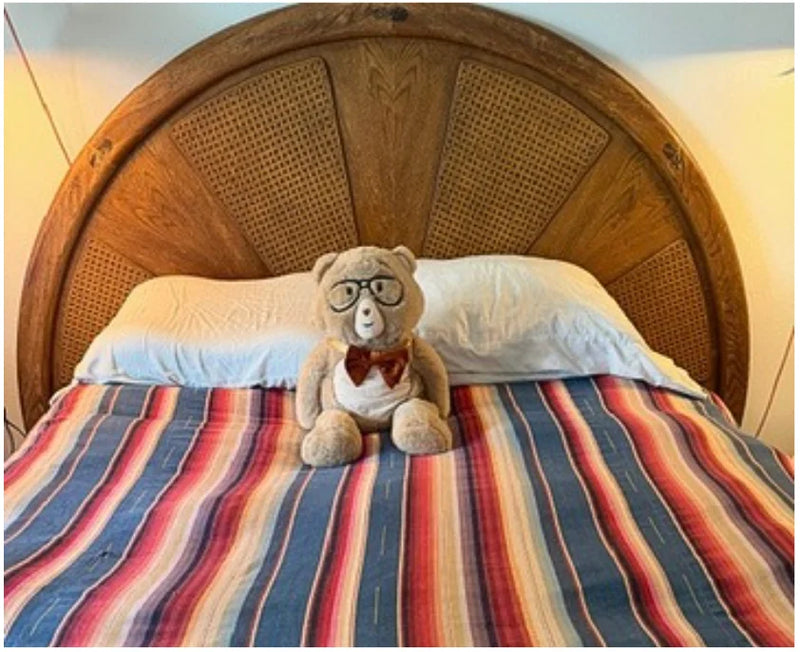 Percy bear bear wearing glasses and a bow tie sitting on a bed with striped bedding and a wooden headboard.