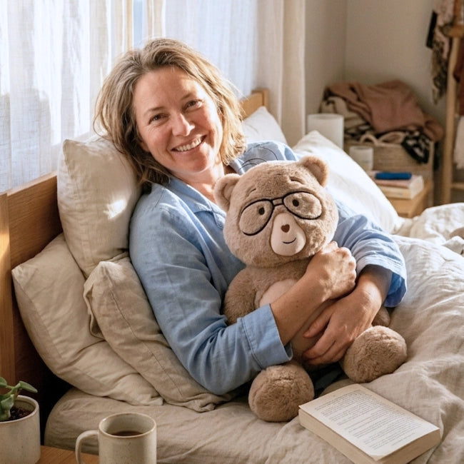 Woman in bed holding a percy bear with glasses, surrounded by pillows and a book.