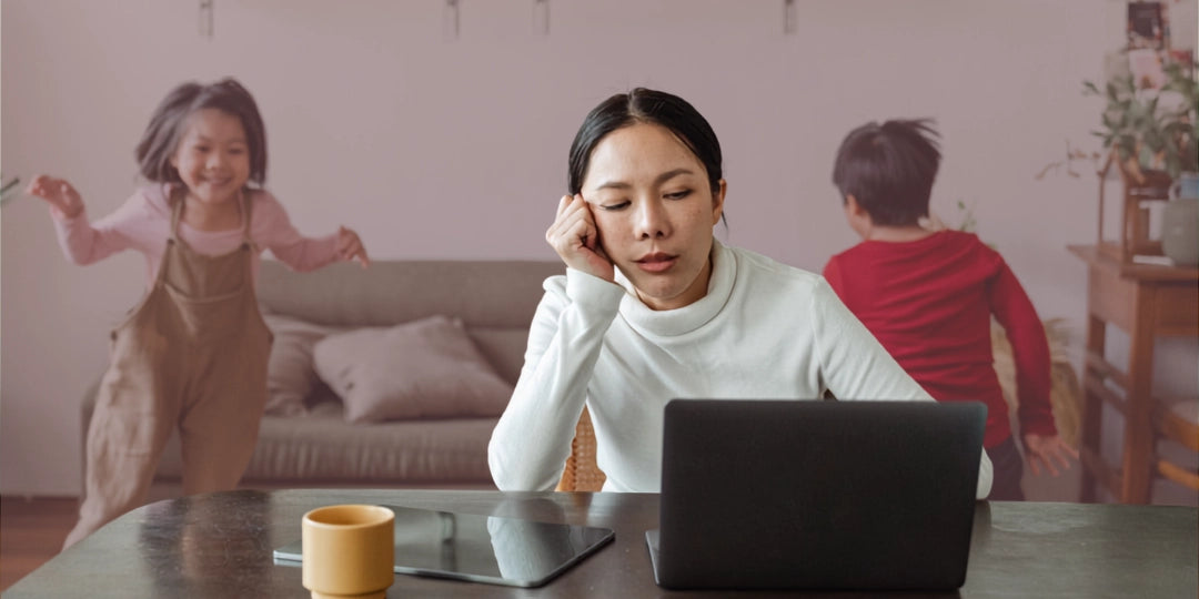 Woman sitting at a table with a laptop, children playing in the background feeling tired