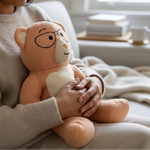 Woman holding a plush Penny weighted teddy bear with glasses in a cozy living room.