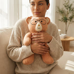 Person holding a plush Penny weighted teddy bear with glasses in a cozy living room.